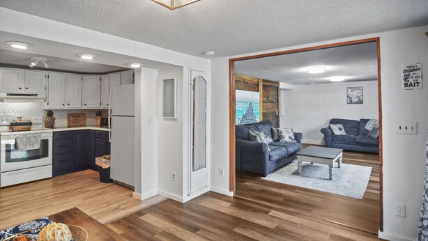 Une cuisine moderne avec des meubles blancs et un plancher en bois, un salon avec un canapé bleu et une table basse, et une vue sur l’océan par une porte.