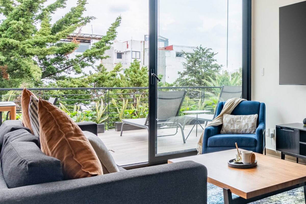 A modern living room with a sofa, armchair, and coffee table, large sliding glass doors leading to a balcony with outdoor furniture and plants.