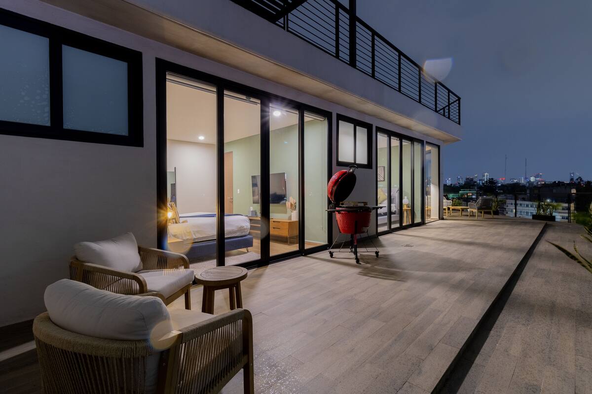 A modern outdoor patio with wicker furniture, a grill, and a glass-enclosed room with a bed and a desk.