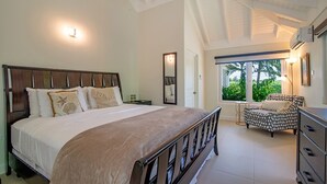 Een slaapkamer met een groot bed, een nachtkastje, een dressoir en een stoel. Er is een raam met uitzicht op groen buiten.
