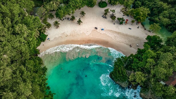Luchtbeeld van een strand met helder turquoise water, omgeven door weelderige groene begroeiing.