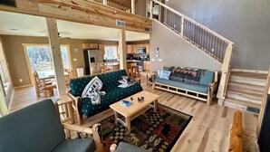 A spacious living area with a high ceiling, wooden beams, a staircase, a sofa, a coffee table, and a kitchen in the background.