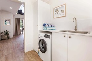A compact kitchen with a washing machine, a sink, and a window with curtains. - Villa 1 - Whitesands (Quindalup)