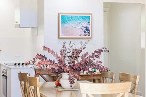 A dining area with a table, chairs, a vase with flowers, a painting on the wall, and a kitchen in the background. - Villa 1 - Whitesands (Quindalup)