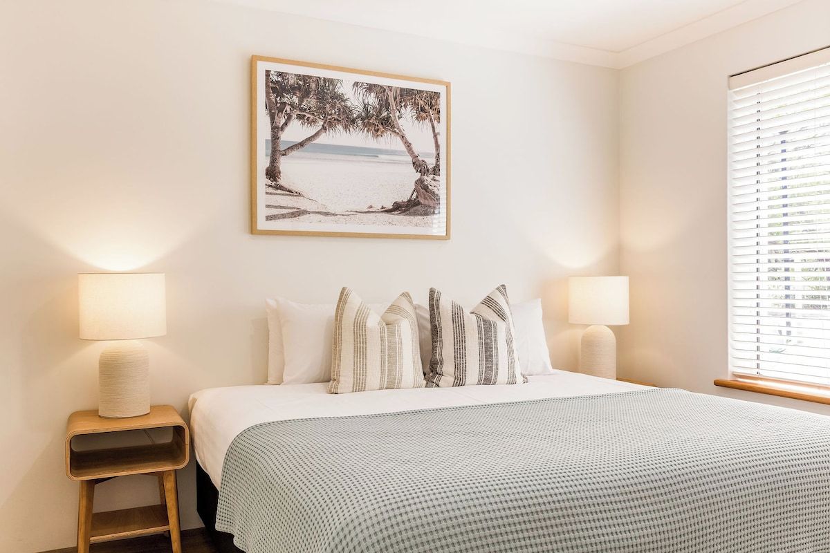 A bedroom with a bed, two bedside lamps, a framed picture on the wall, and a window with blinds.
