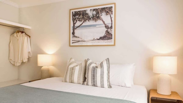 Una habitación de hotel con cama, perchero, un cuadro de una playa y dos lámparas de noche.