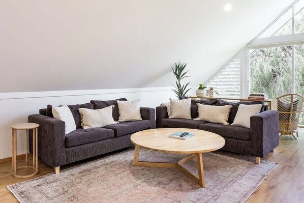 A modern living room with two dark sofas, a round wooden coffee table, a small side table, and a large window with a view of trees.