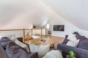 Un appartement moderne de style « loft », doté d’un plafond haut, d’un escalier en bois, d’un espace de vie avec un ensemble de canapés, une table basse et un téléviseur à écran plat fixé au mur.