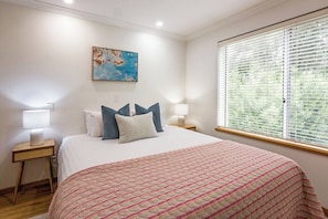 Une chambre à coucher comprenant un lit, deux tables de chevet, une fenêtre avec des stores et un tableau accroché au mur.