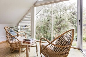 Une pièce dotée d’une grande porte-fenêtre coulissante donnant sur un jardin, de meubles en osier et d’une petite table sur laquelle sont posés des livres.