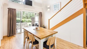 A dining area with a wooden table and chairs, a view through a glass door to an outdoor space, and a staircase with wooden railings. - Whitesands Villa 3 (Quindalup)