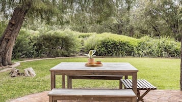 A wooden picnic table with a bench, surrounded by a grassy area and dense shrubbery.