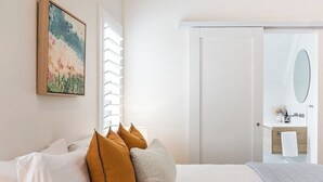 Quarto de hotel com cama, almofadas, um quadro, janela com persianas e banheiro com espelho e pia.