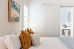 Een hotelkamer met een bed, kussens, een schilderij, een raam met zonwering en een badkamer met spiegel en wastafel.