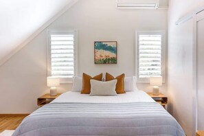 Une chambre avec un plafond en pente, deux tables de chevet avec des lampes, un lit recouvert d’une couverture à rayures et un tableau accroché au mur.