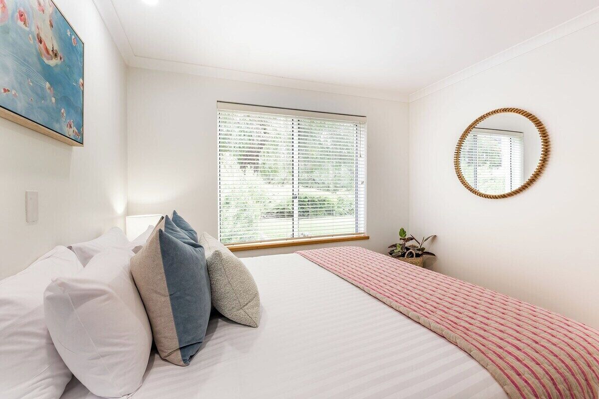Une chambre à coucher avec un lit, des oreillers, une fenêtre avec des stores, un miroir et un tableau accroché au mur.