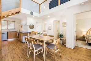 A spacious dining area with a wooden table and chairs, a kitchen in the background, and a staircase leading to an upper level.