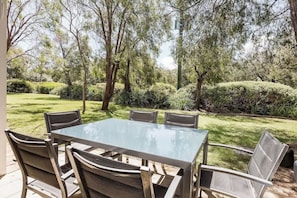 An outdoor patio with a glass-top table and six chairs, surrounded by trees and a well-maintained lawn.