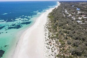 An aerial view of a coastal area with a sandy beach, clear blue waters, and a dense vegetation-covered hillside.