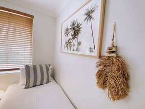Un dormitorio con cama, una ventana con persianas, un cuadro enmarcado con palmeras y un adorno colgado en la pared.