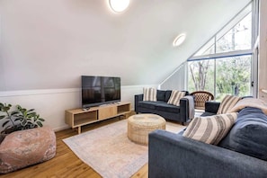 Un salon moderne avec un téléviseur à écran plat, des canapés sombres, un meuble TV en bois, une grande fenêtre et une plante en pot.