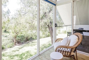 Une pièce dotée de grandes portes vitrées, d’un fauteuil en osier, d’une petite table ronde et offrant une vue sur les arbres et la verdure environnante.