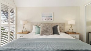 Un dormitorio con una cama grande, dos mesitas de noche con lámparas, un cuadro enmarcado en la pared y una ventana con persianas.