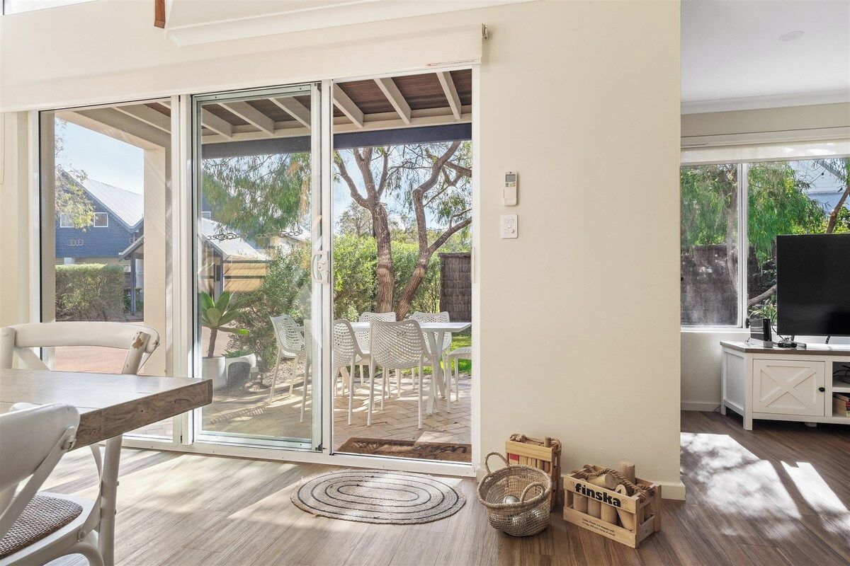 A room with a sliding glass door leading to an outdoor patio with a table and chairs, a television on a stand, and a wooden floor.