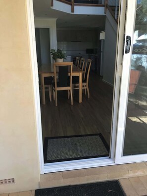 Une porte ouverte donne sur un espace repas avec une table et des chaises, une plante en pot, et une vue sur une cuisine et un niveau supérieur.