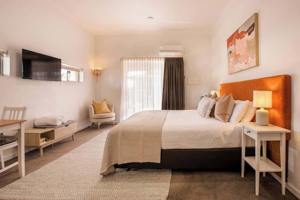 A hotel room with a bed, bedside table, chair, TV, air conditioning unit, and a painting on the wall.
