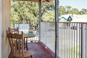 A balcony with a table and chairs, a white railing, and a view of a parking area and trees.