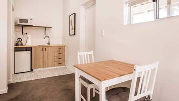 A compact kitchen with a microwave, kettle, and small refrigerator. There is a wooden table with chairs and a framed picture on the wall.