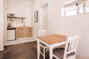 A compact kitchen with a microwave, kettle, and small refrigerator. There is a wooden table with chairs and a framed picture on the wall.