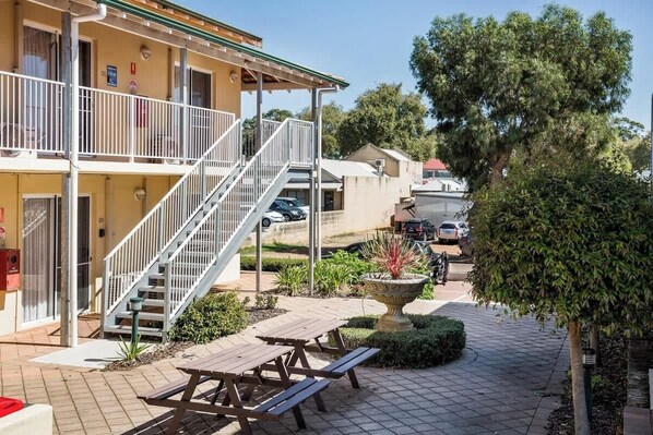 Un bâtiment doté d’un balcon, d’un escalier et d’un espace jardin agrémenté d’une fontaine et de tables de pique-nique.