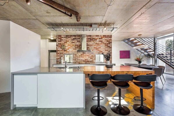 Una cocina moderna con pared de ladrillo, una isla central con taburetes de bar y una escalera que sube a un nivel superior.