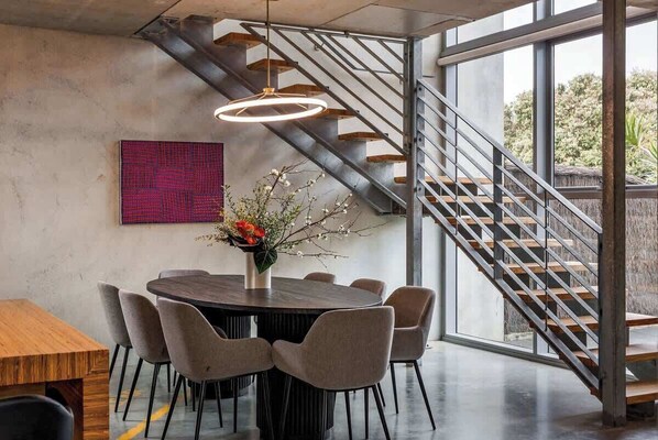 A modern dining area with a round table, chairs, a vase with flowers, a staircase, and a large window.