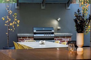 An outdoor grill area with a brick wall, a table with chairs, and a potted plant. - Djurapin—Sophisticated Coastal Haven with Jacuzzi (Dunsborough)