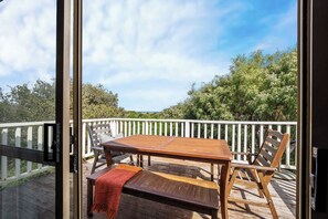 Una terraza exterior con una mesa y sillas de madera, barandilla blanca y vistas a árboles y cielo.