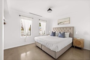 A bedroom with a large bed, two windows with curtains, a headboard, a bedside table with a lamp, and a painting on the wall.