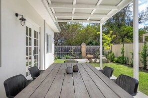 Eine überdachte Terrasse mit einem Holztisch und Stühlen, einer weißen Außenwand und einem umzäunten Garten.