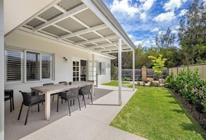 Eine überdachte Terrasse mit Tisch und Stühlen, eine weiße Außenwand und ein gepflegter Rasen.
