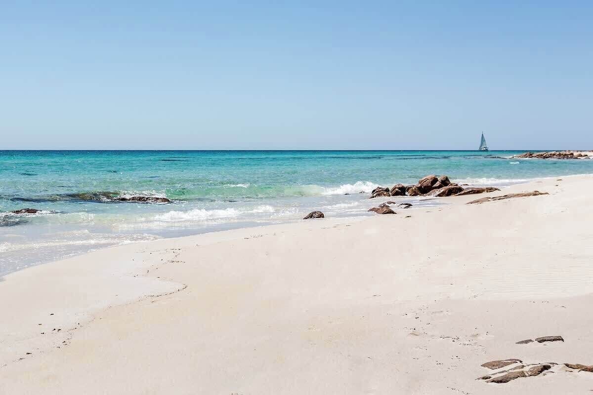 Une plage bordée d’une eau turquoise et limpide, un voilier à l’horizon et des rochers éparpillés le long du rivage.