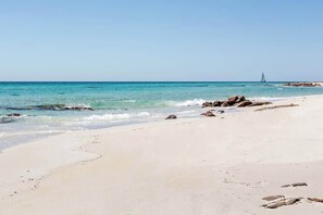 Een strand met helder turquoise water, een zeilboot in de verte en verspreide rotsen langs de kust.