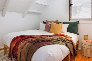 A bedroom with a bed, pillows, a blanket, a window with blinds, and a small bedside table.