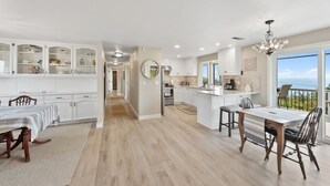 A spacious kitchen with white cabinetry, a dining area with a wooden table, and a view of the ocean through large windows. - Easy Beach Access, Spacious Decks & Striking Ocean Views - Harris Beach Overlook (Brookings)