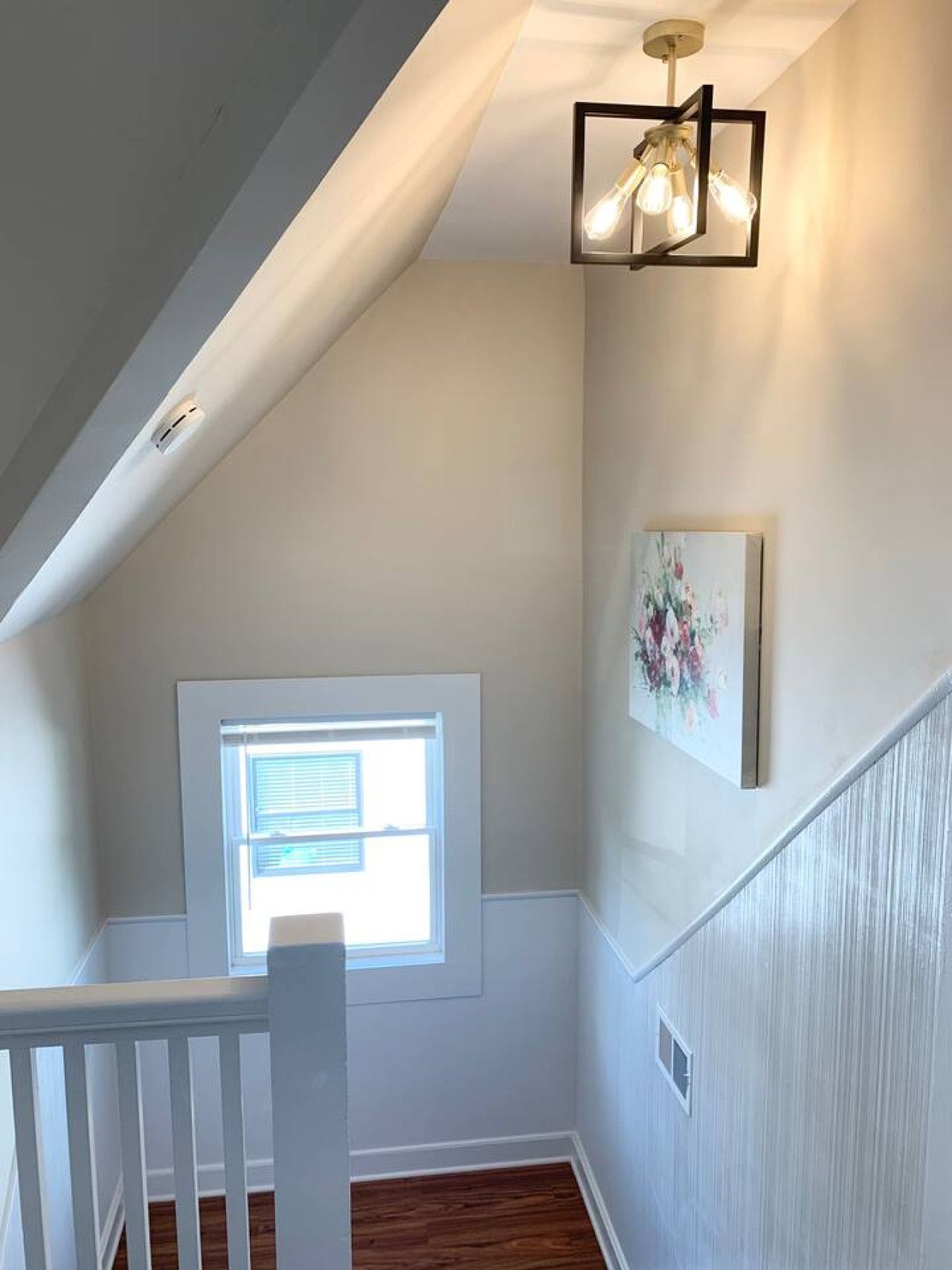 A room with a sloped ceiling, a window with blinds, a white railing, and a wall-mounted light fixture.