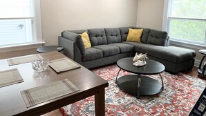 Un salon moderne avec un canapé d’angle gris, une table basse ronde et une table à manger agrémentée de sets de table.