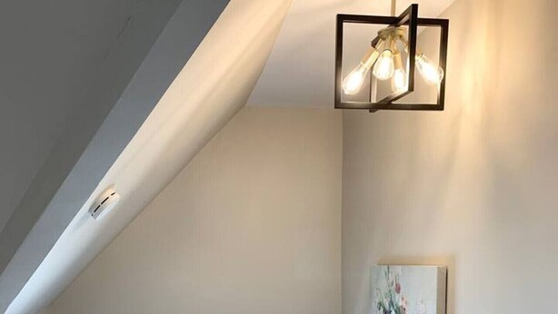 Une pièce avec un plafond en pente, une fenêtre avec des stores, une rambarde blanche et un luminaire fixé au mur.