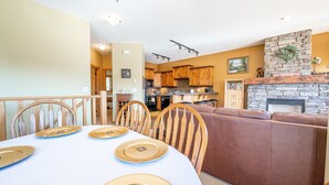 A dining area with a table set for four, a brown sofa, and a kitchen in the background. - Bear's Paw Chalet - Amazing Location & Ski In/out (Beaverdell)