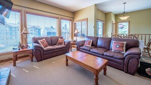 A spacious living room with a brown leather sofa set, a wooden coffee table, and a lamp on a side table. Large windows with blinds allow natural light to fill the room. - Bear's Paw Chalet - Amazing Location & Ski In/out (Beaverdell)